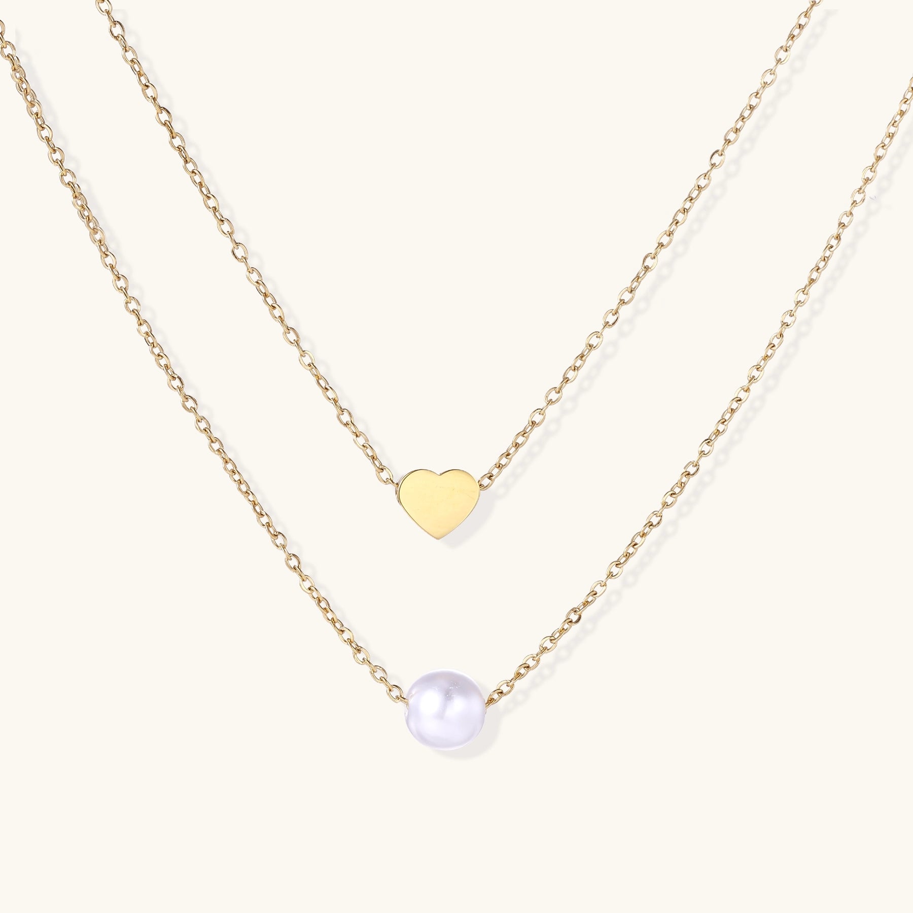 Two delicate gold-plated chains layered together, one with small heart pendant, another with cream pearl bead