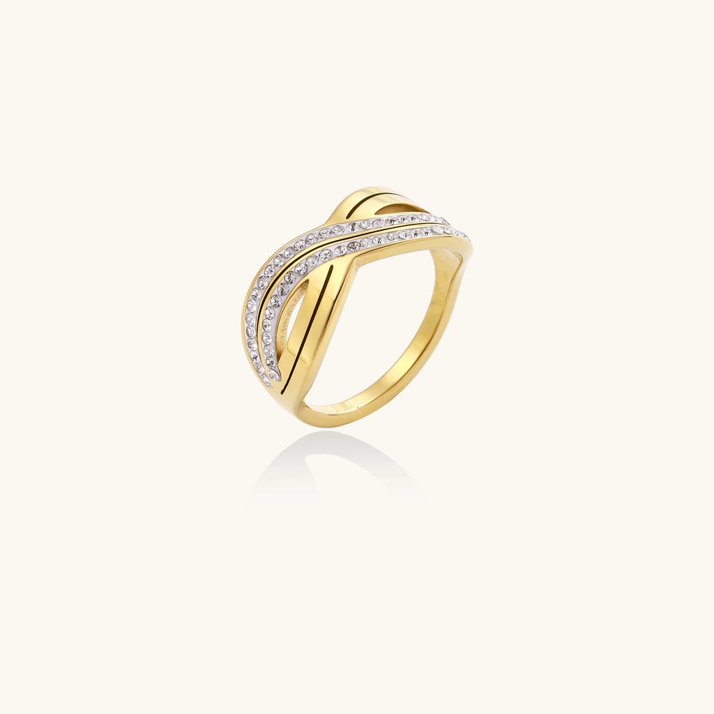 Gold-plated stainless steel ring with crossed infinity bands adorned with sparkling rhinestones against cream background.