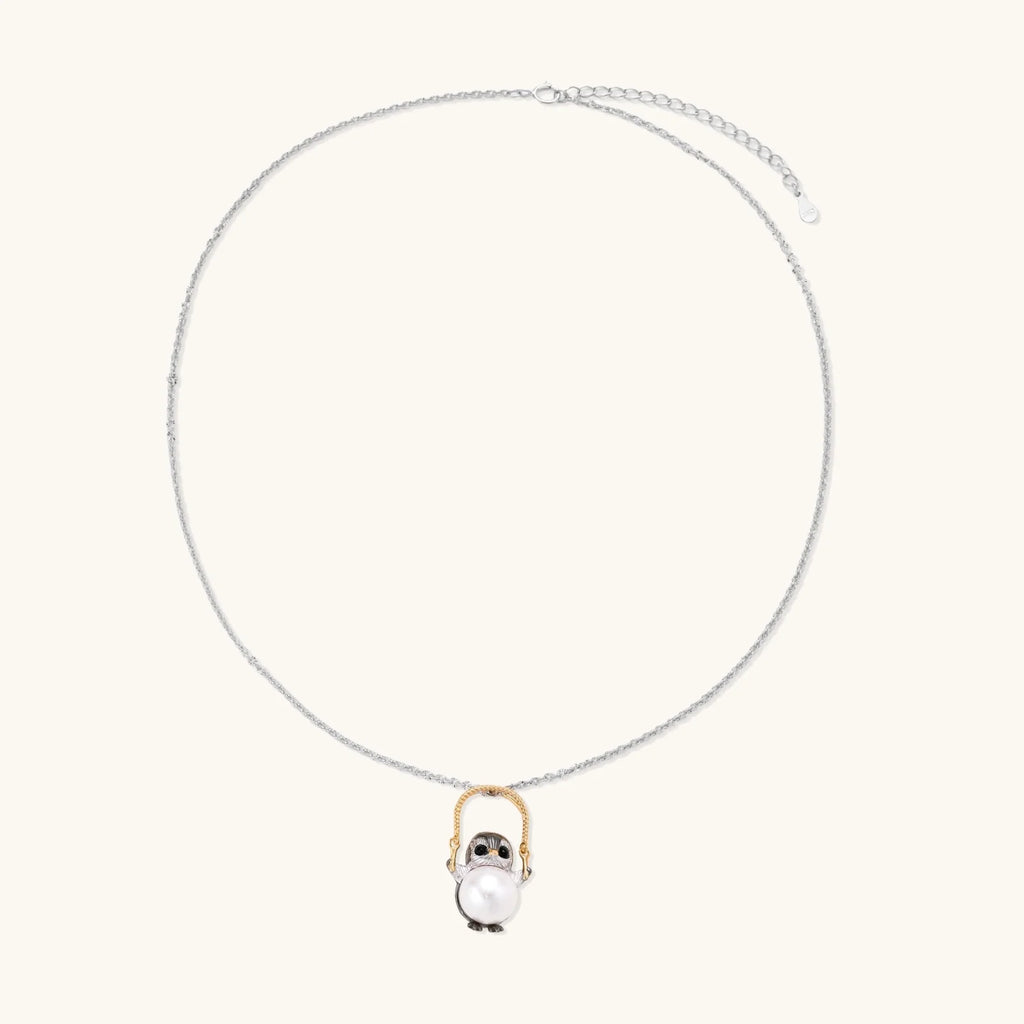 Whimsical owl pendant holding a white pearl on a gold swing, rhodium-plated silver chain on cream background.