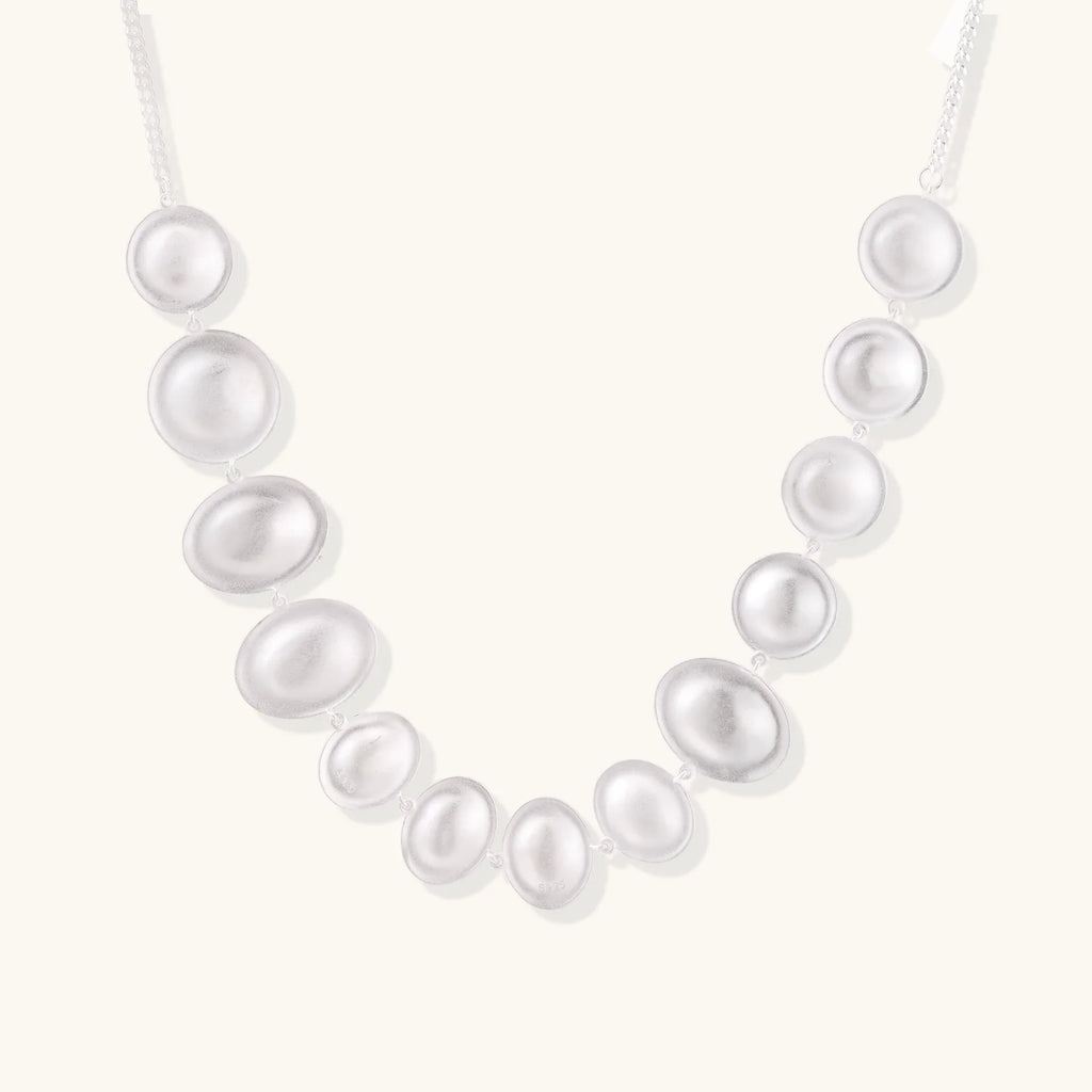 Graduated pearl-finish silver discs forming an organic bubble bib necklace on delicate chain against cream background.